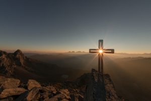 Photo of a cross on top of a mountain highlighted by the rising sun